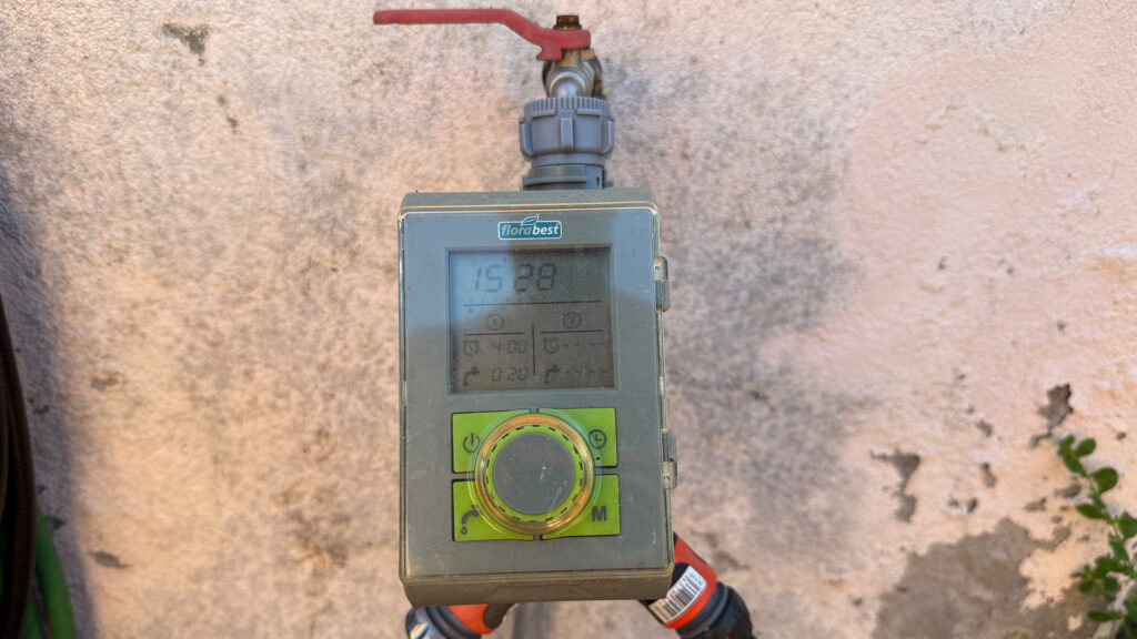 Ein einfacher Bewässerungsautomat mit Digitalanzeige mit zwei Auslässen an einem Wasserhahn vor einer leicht fleckigen, verputzten Mauer. 