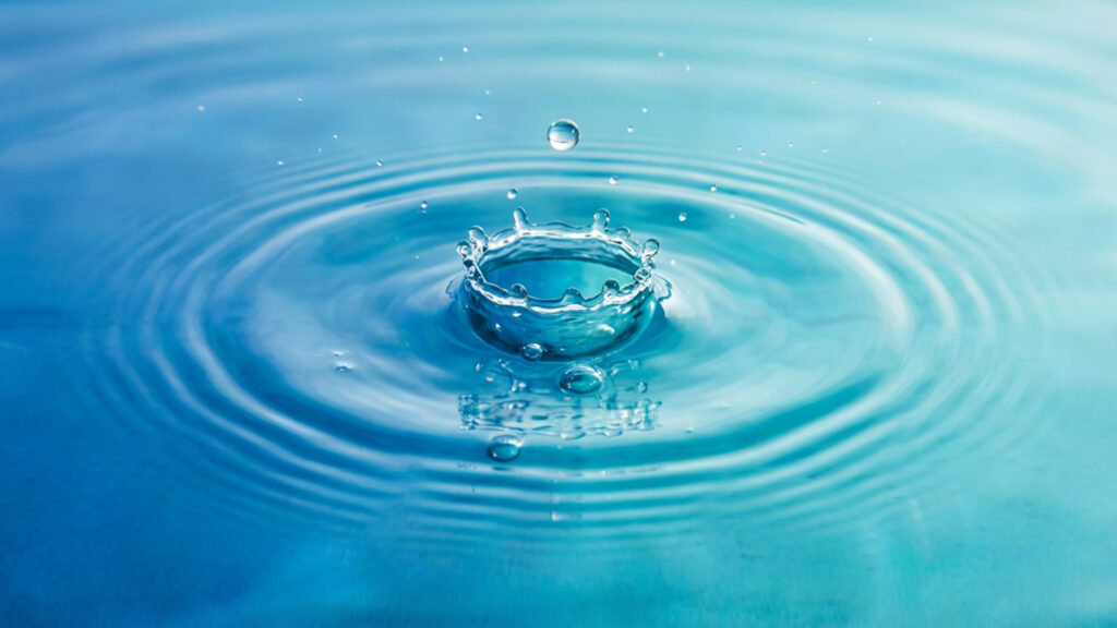 Ein Wassertropfen, fällt ins Wasser. Um die Stelle herum sieht das Wasser aus wie eine kleine Krone. Das Wasser ist blau bis türkis und schlägt ein paar kleine Wellen.