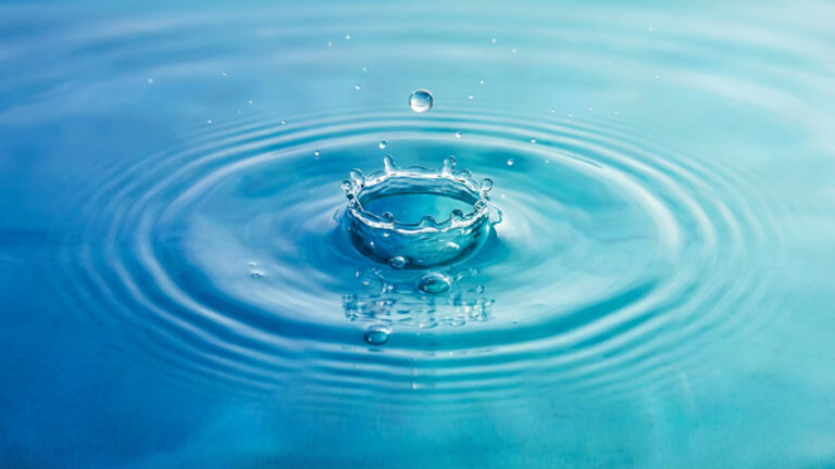 Ein Wassertropfen, fällt ins Wasser. Um die Stelle herum sieht das Wasser aus wie eine kleine Krone. Das Wasser ist blau bis türkis und schlägt ein paar kleine Wellen.